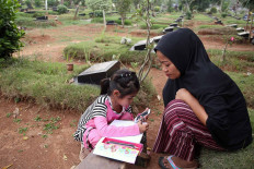 Adinda Rahma Barokah, 7, squats near a tombstone while doing her school assignment in Padurenan cemetery in Bekasi, West Java, on Oct. 15. She read the questions on her mother&rsquo;s phone and wrote down the answers in her notebook. As her school -- SD Wanarasi 15 state elementary school -- is closed due to the outbreak, Adinda has been studying in the cemetery where her mother sells snacks and drinks. JP/P.J.Leo